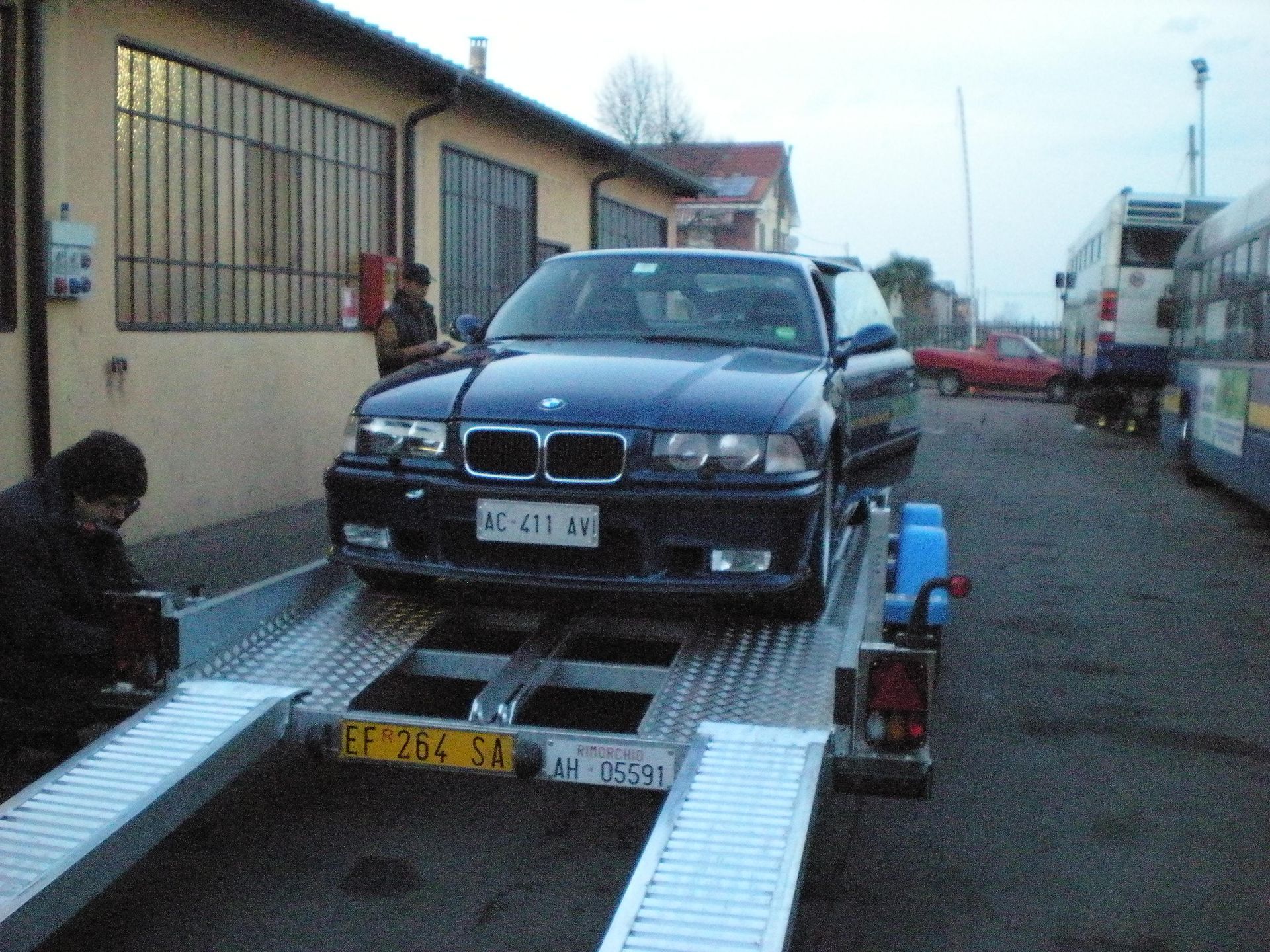 Una BMW blu scuro su un rimorchio caricata da due persone all'esterno di un edificio.