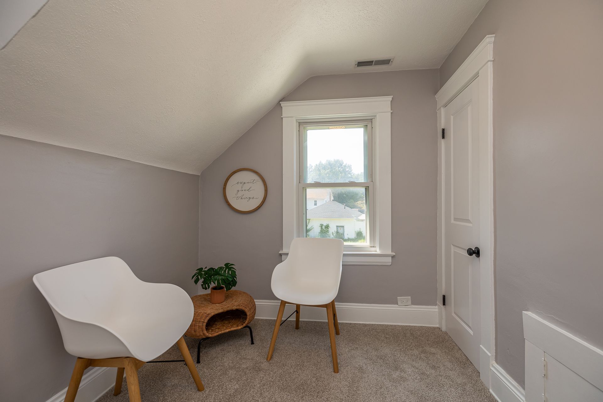 Cozy attic space with two white chairs, small table, and a window. Light gray walls, beige carpet.