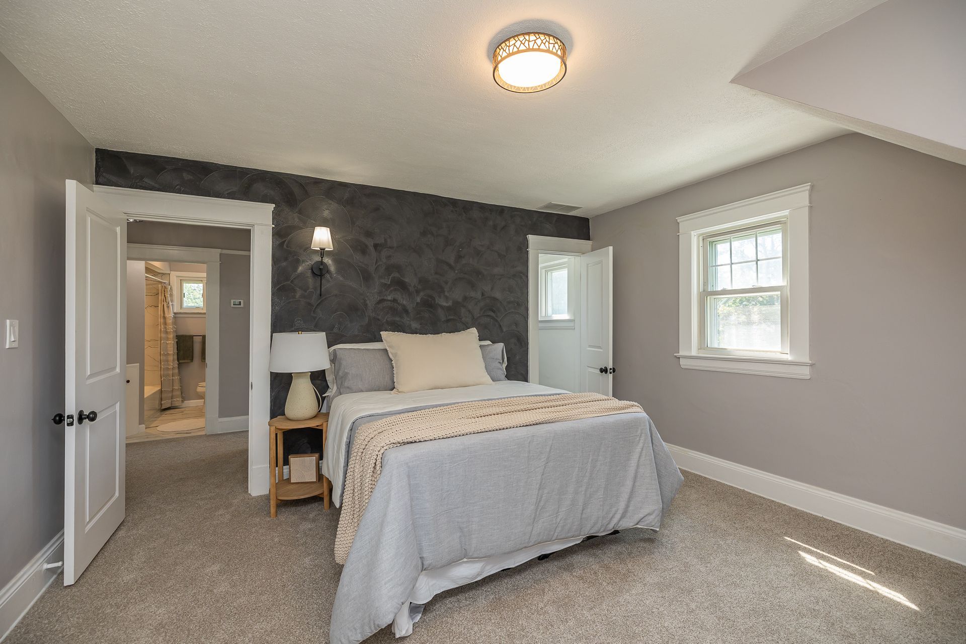 Bedroom with bed, nightstand, and window. Gray walls and carpet. Door to bathroom open.