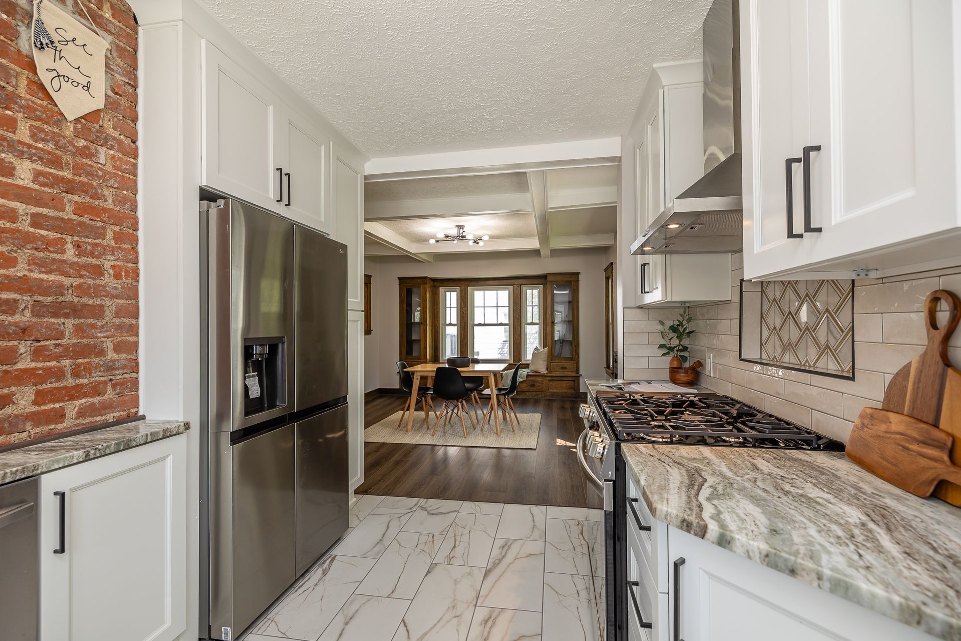 Galley kitchen with stainless steel appliances, white cabinets, brick wall, and view into a dining area.