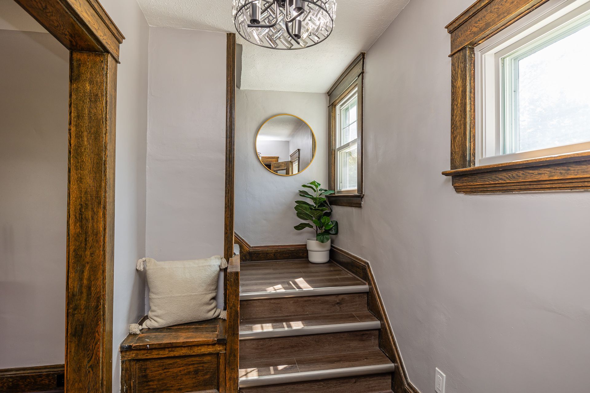 Stairway with wood trim, a round mirror, and a small potted plant. Gray walls, bench with pillow, and a bright window.
