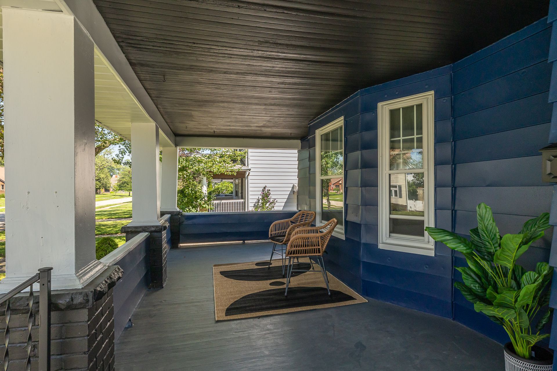 Covered porch with blue siding, white trim, and a rug with chairs, and potted plant.