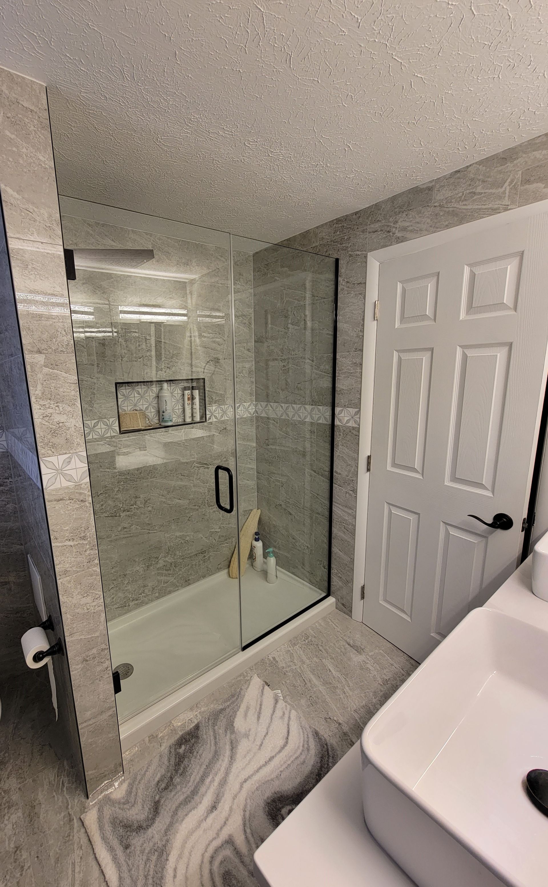 Modern bathroom with gray tiled shower and white door. White sink and gray swirl rug.