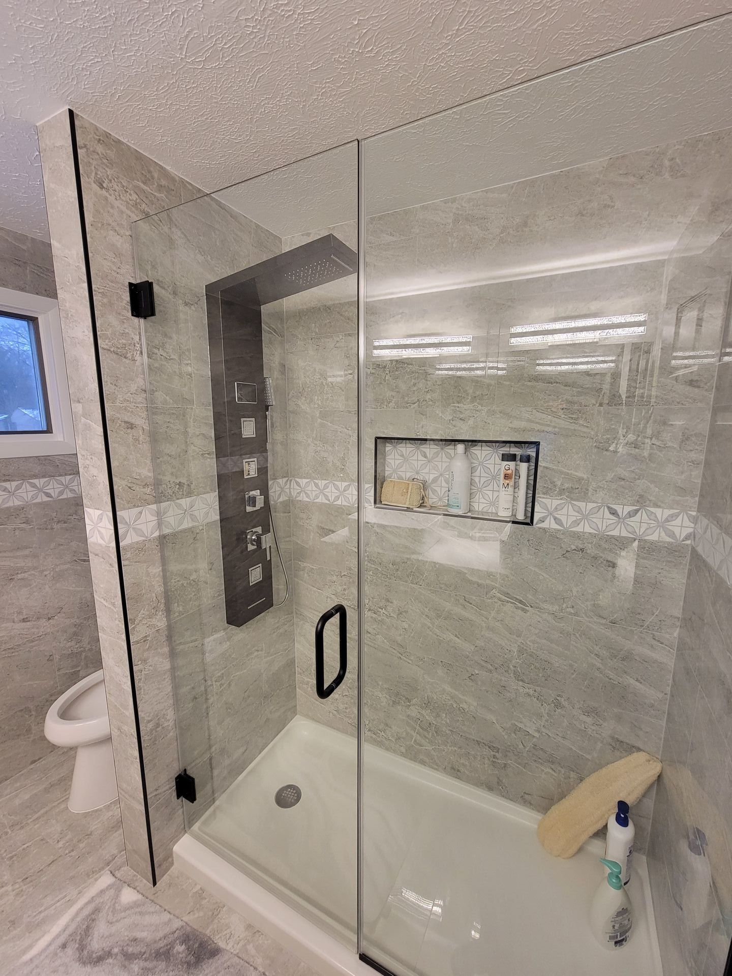 Modern shower with glass door and gray tiled walls, recessed shelf, and multi-jet shower system.