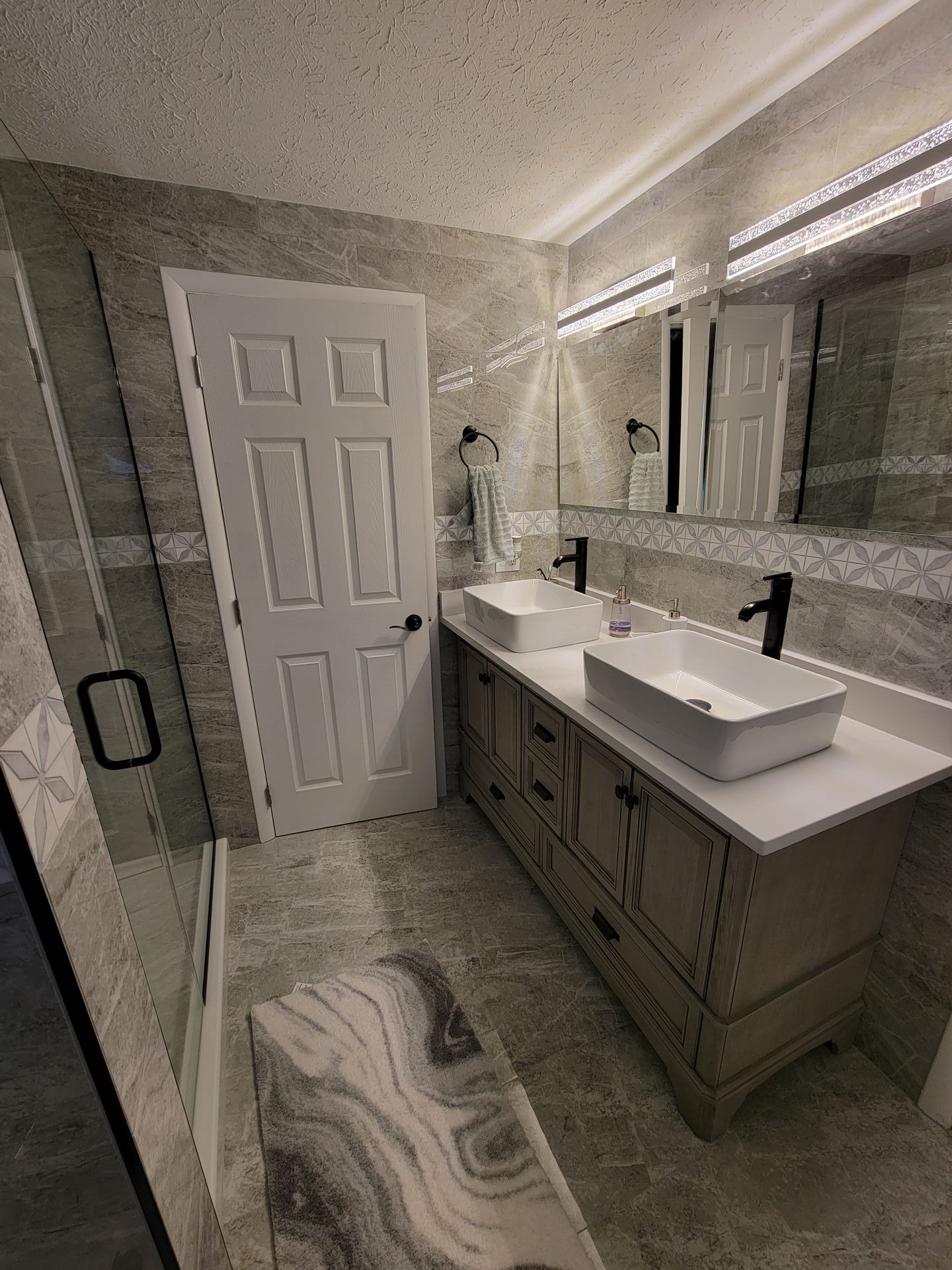Bathroom with gray tiled walls, a white door, double vanity with rectangular sinks, and a glass shower.