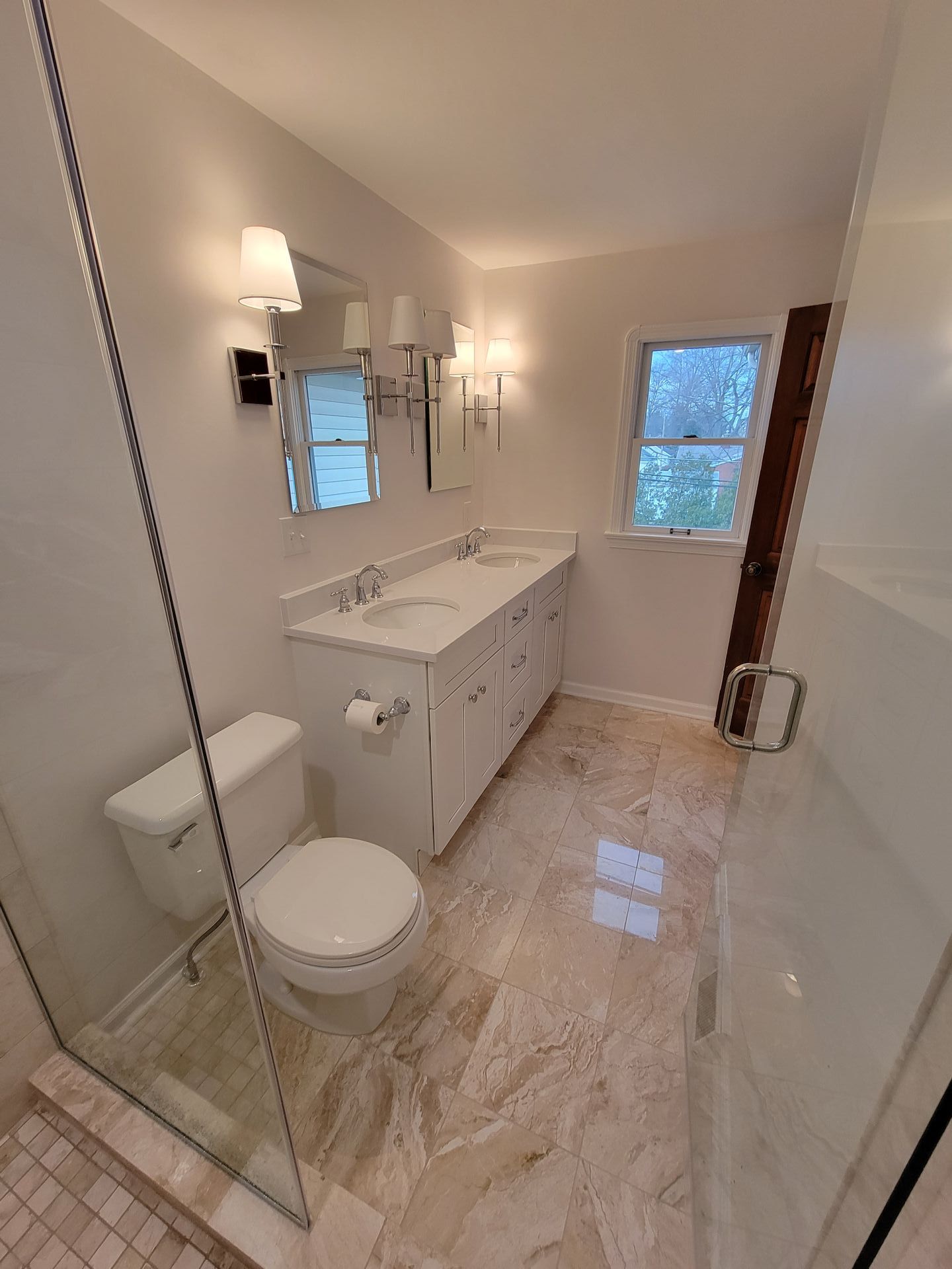 Bright bathroom with a white double vanity, toilet, and marble-look floor. Glass shower and window visible.