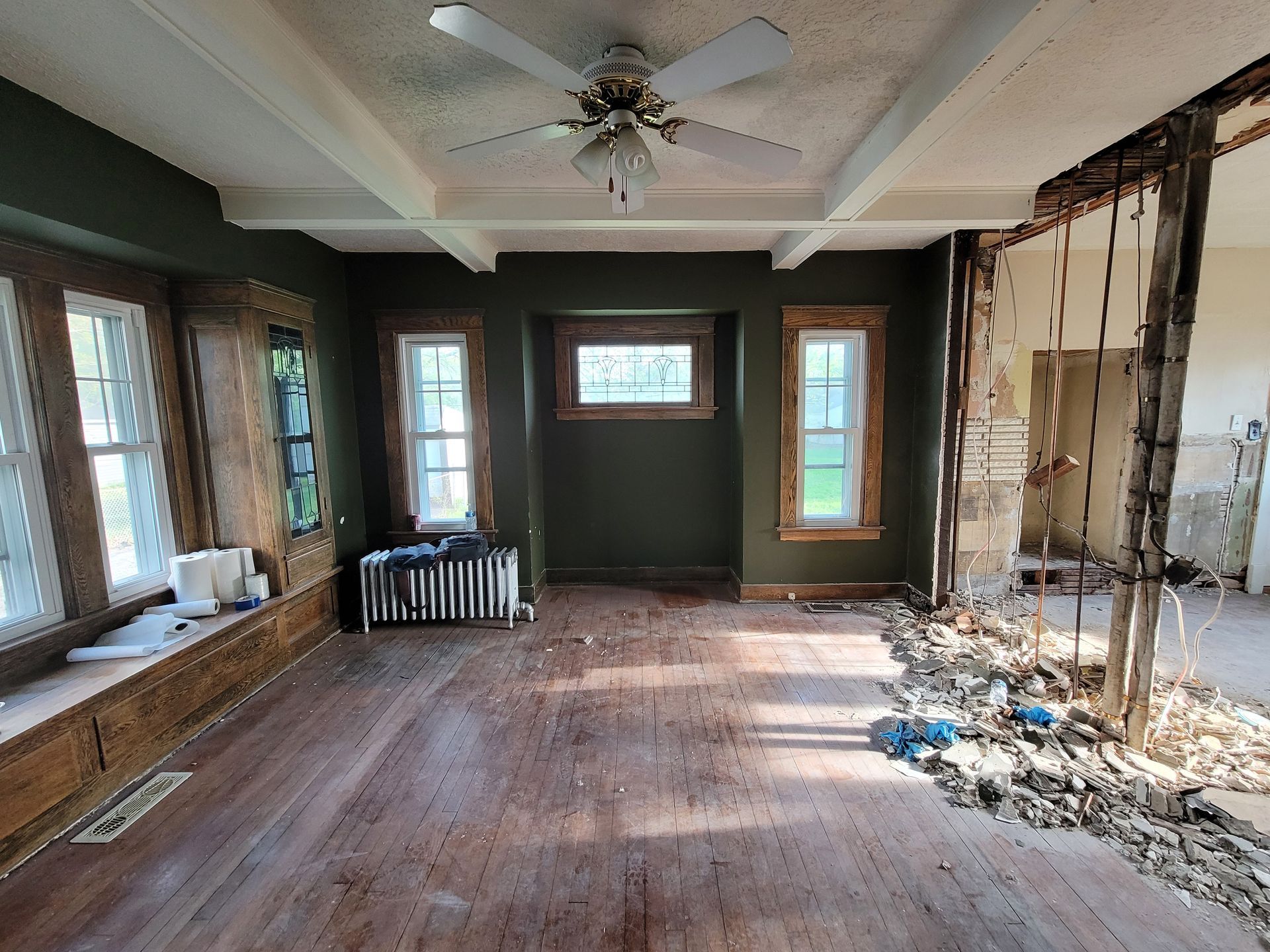 Room undergoing renovation, dark green walls, exposed support beams, hardwood floors, debris on floor.