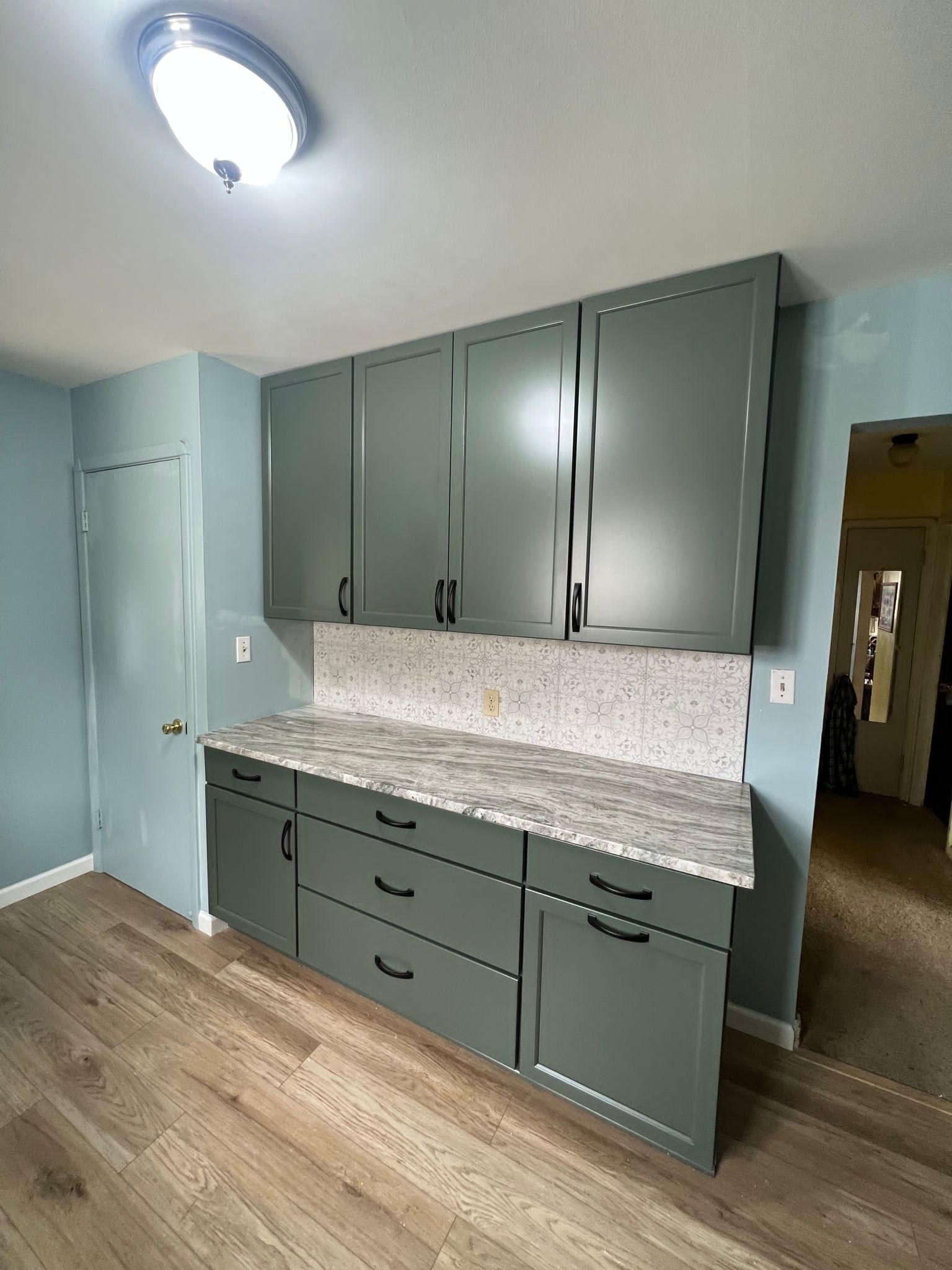 Sage green kitchen cabinets with granite countertop, neutral backsplash, and wood flooring.