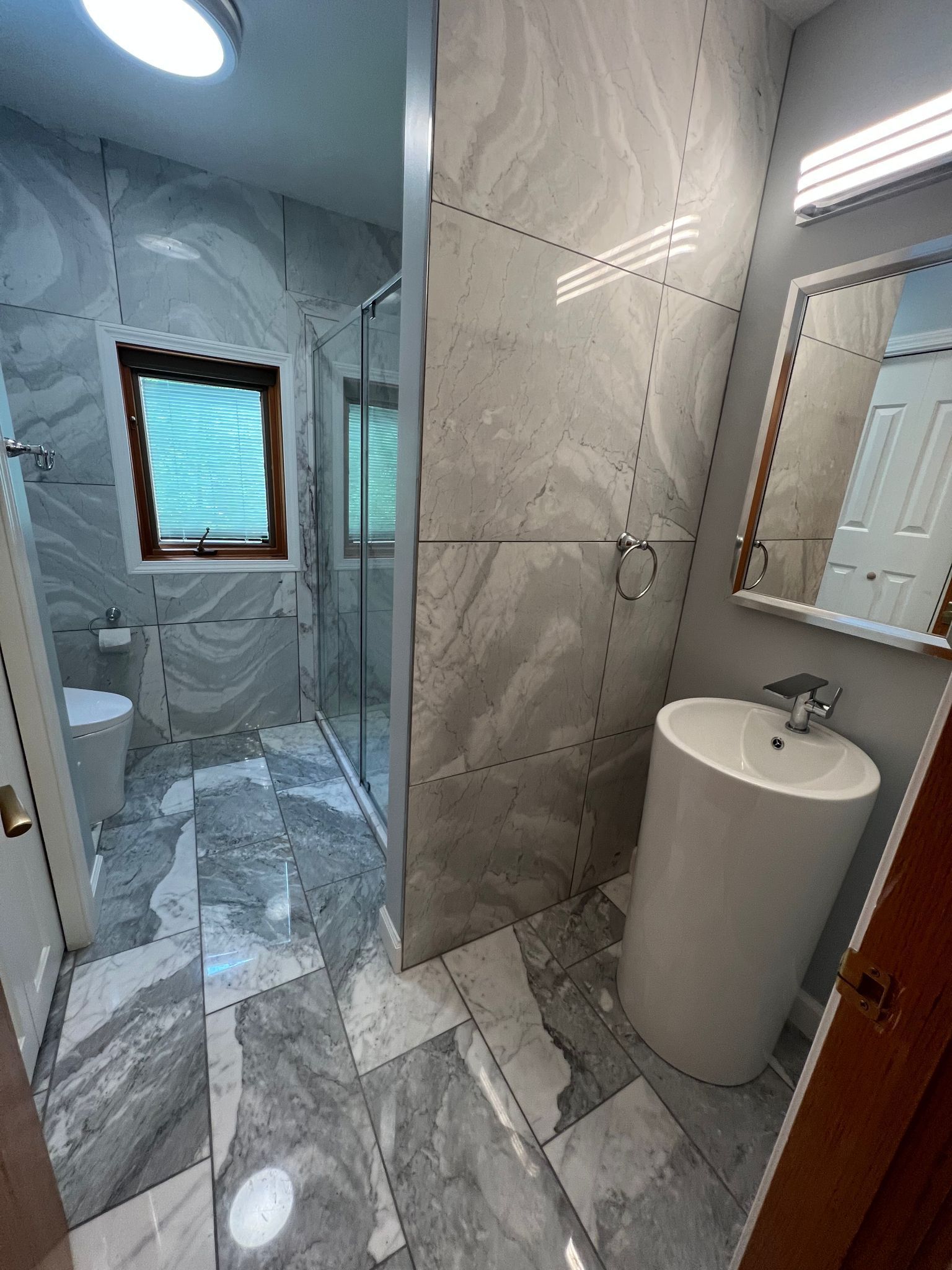 Gray and white tiled bathroom with a pedestal sink, mirror, shower, and toilet.