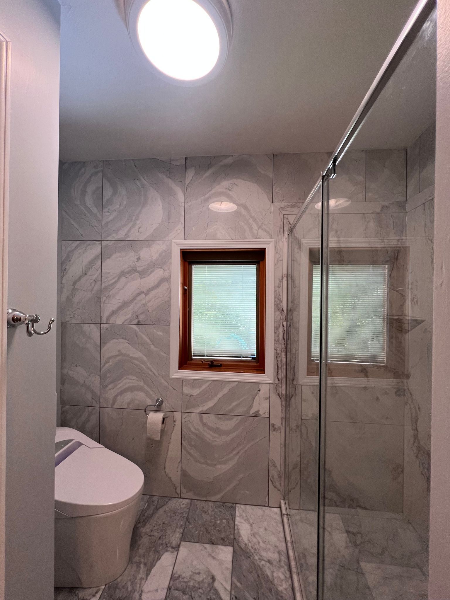 Bathroom with marble tile walls, a toilet, a small window, and a glass shower door.
