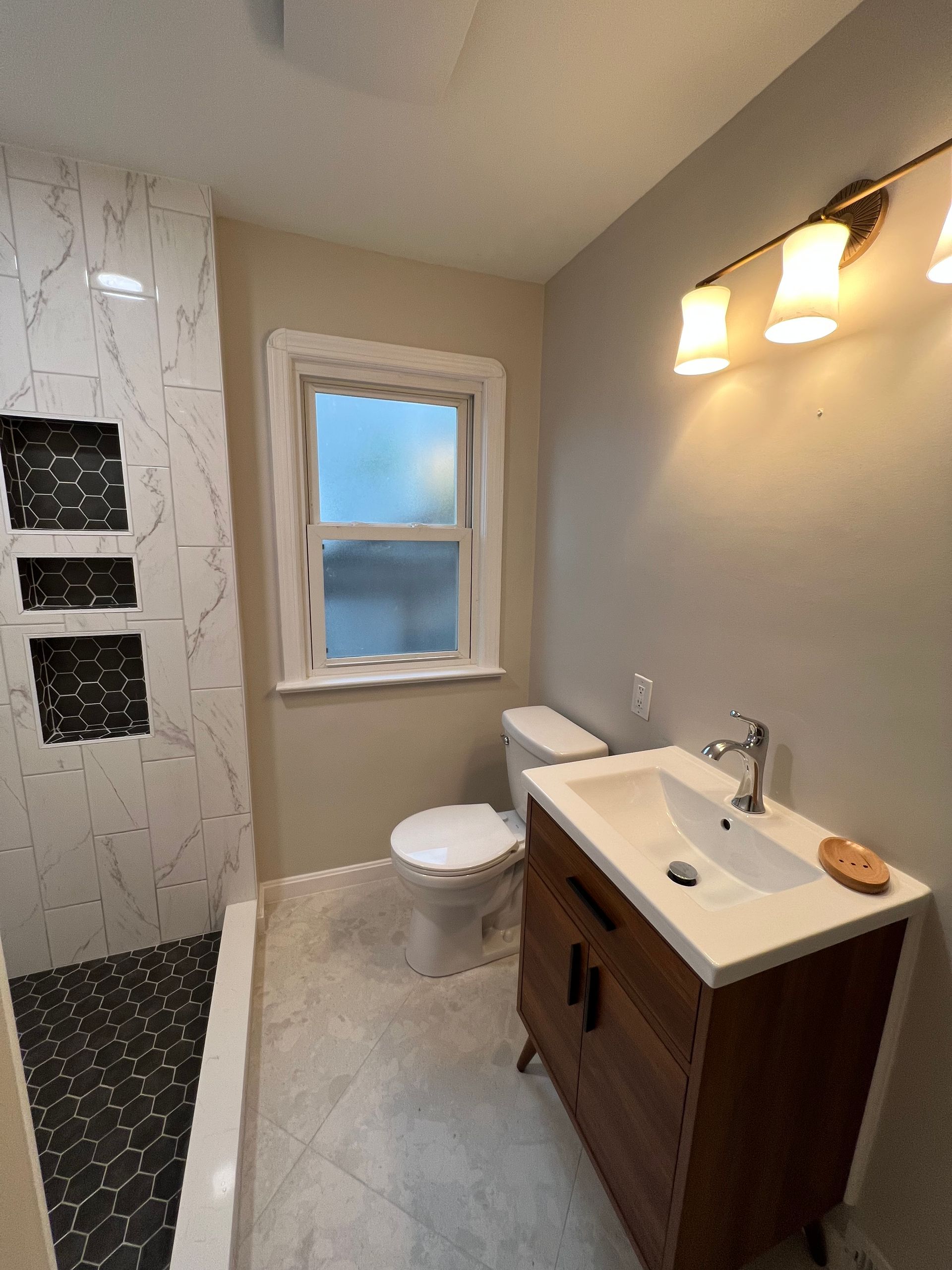 Bathroom with shower, vanity, toilet, and window. Gray walls, white and dark tile, wooden vanity.