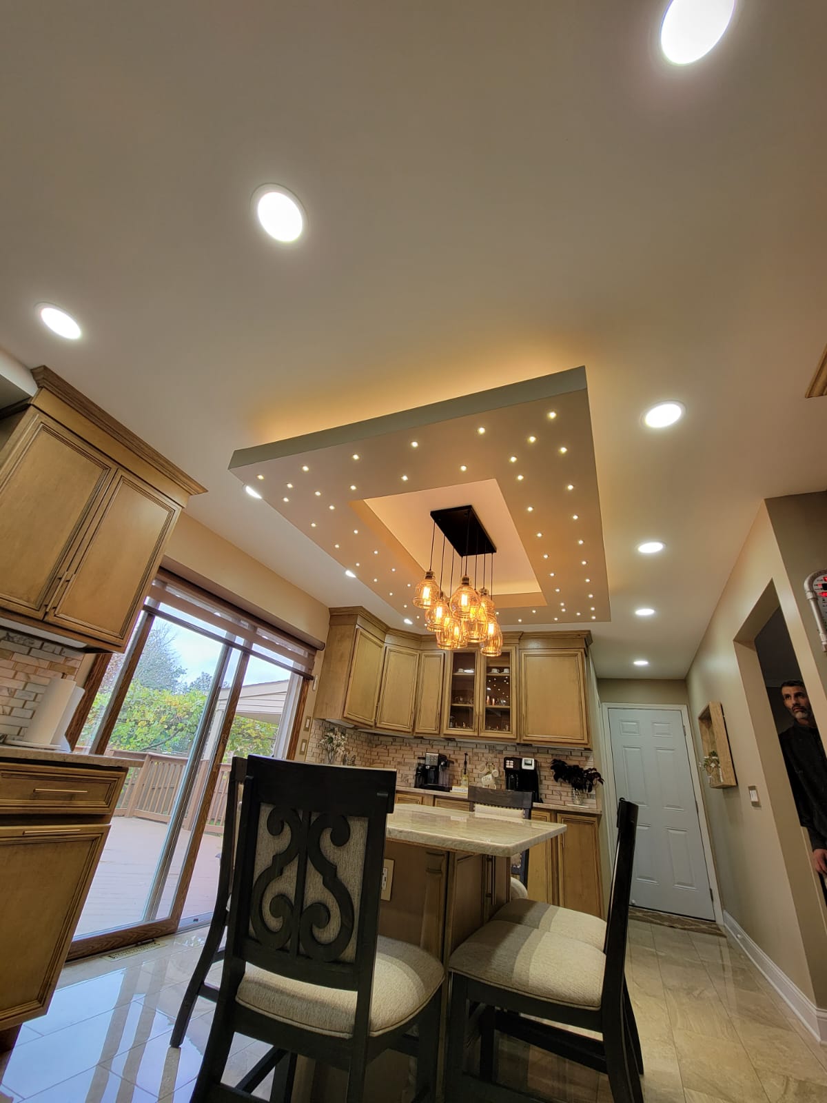Kitchen with a tray ceiling lit by multiple lights and a hanging light fixture. Cabinets and chairs are visible.