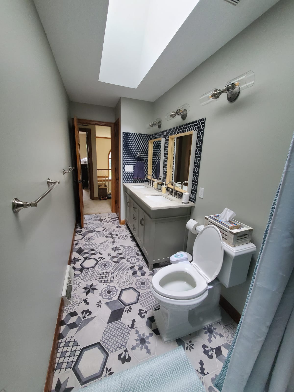 Bathroom with blue, grey, and white patterned floor tiles, double vanity, and a toilet. A window is in the ceiling.
