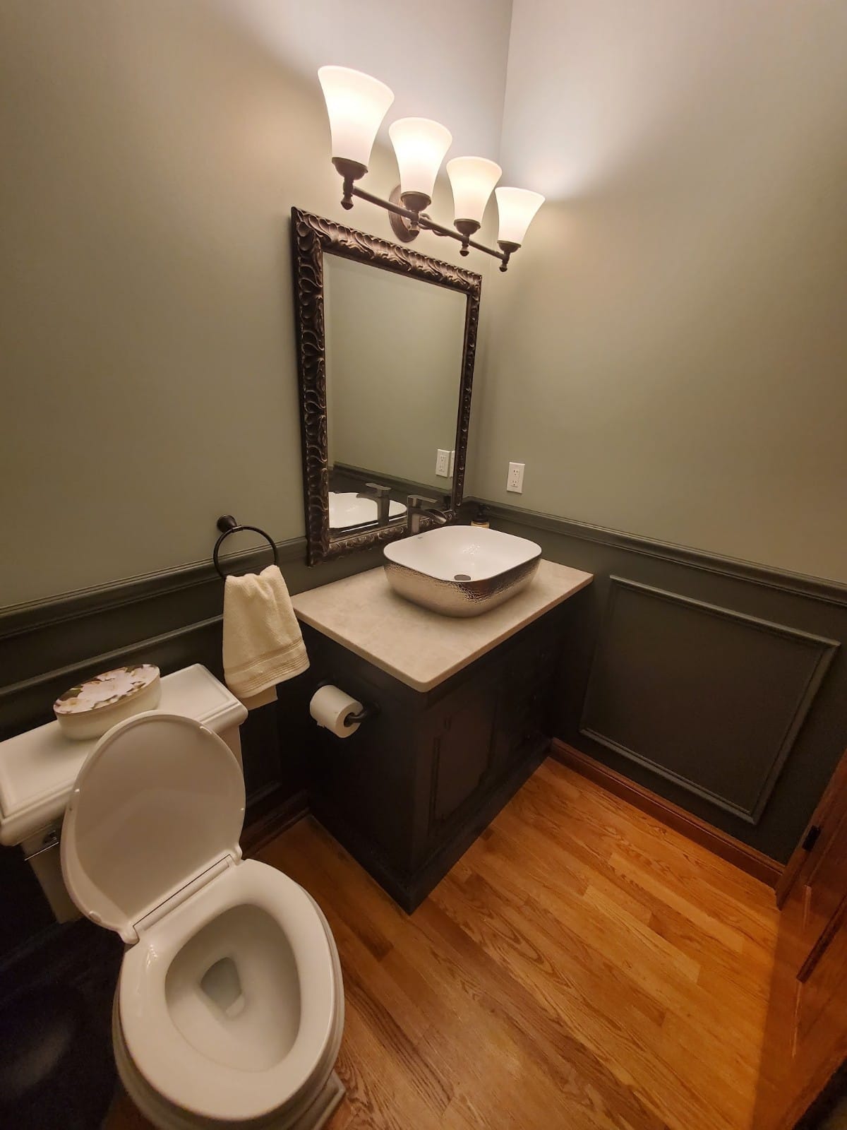 A powder room with a toilet, sink, mirror, and a wall-mounted light fixture. Olive green walls and wood floors.