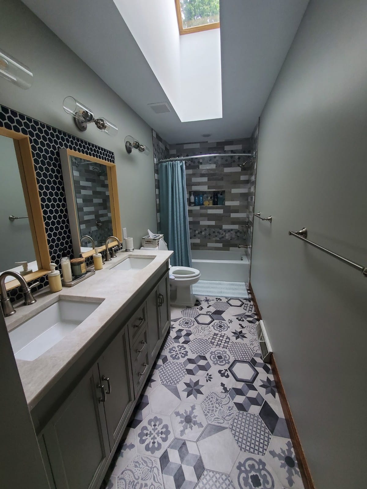 Bathroom with double vanity, patterned floor, shower, and skylight. Light green walls, gray vanity, and blue shower curtain.