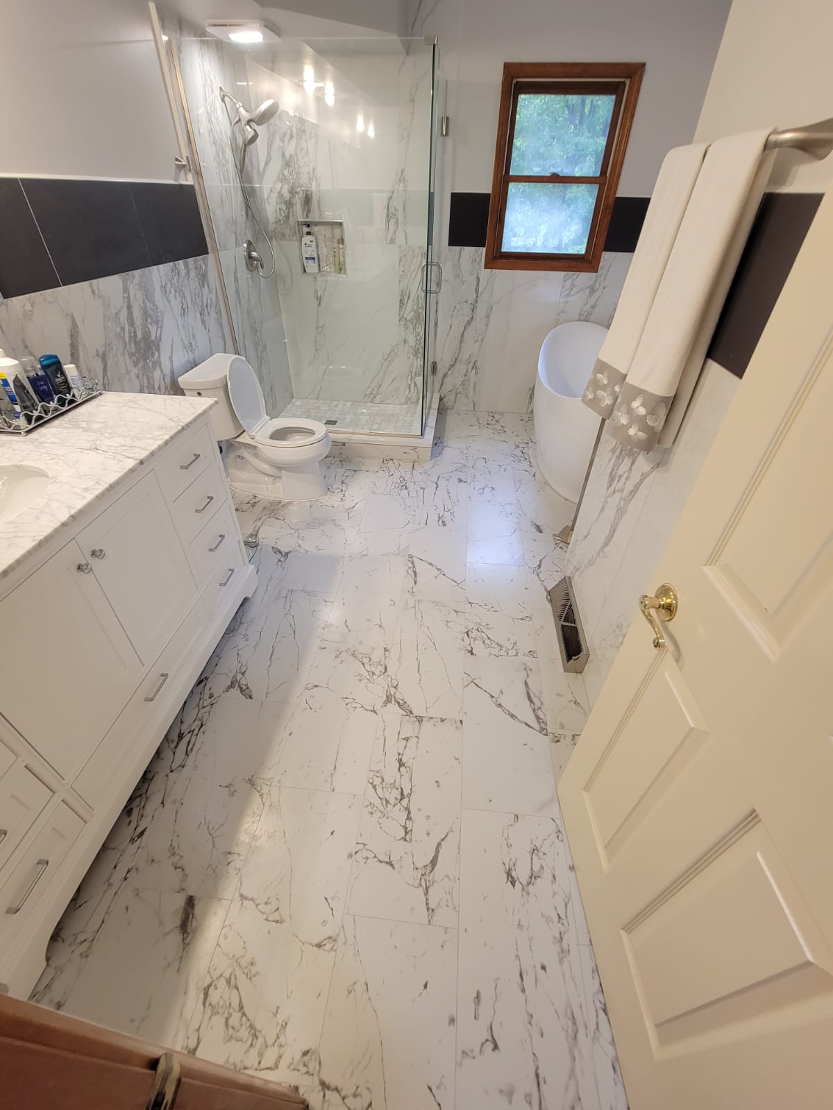 White and marble-tiled bathroom with a glass shower, toilet, bathtub, vanity, and a partially open door.