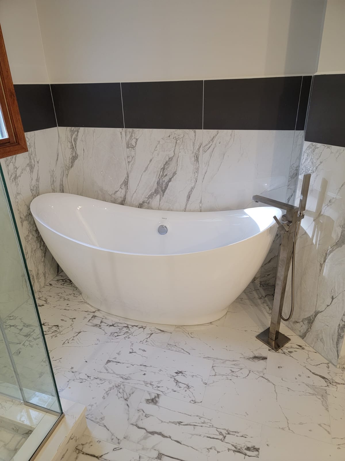 White freestanding bathtub in a bathroom with marble-look tile, dark tile accents, and a floor-mounted faucet.