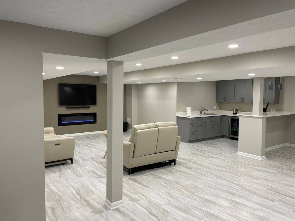 Finished basement with a TV above fireplace, seating, and a kitchenette. Gray walls, light wood-look flooring.
