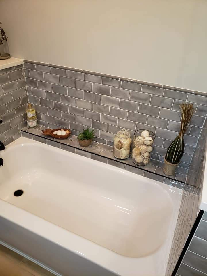 White bathtub with gray tiled backsplash, glass shelf holding candles and decorative items.