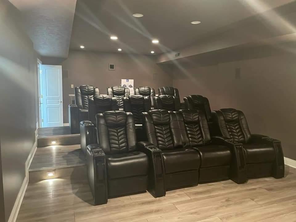 Home theater with tiered seating of black leather recliners; grey walls, and wood-look flooring.