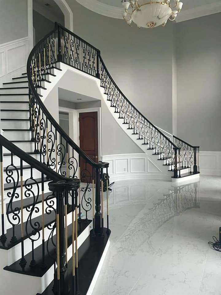 Grand curved staircase with black and gold railing, leading to a second floor landing. White walls and grey flooring.