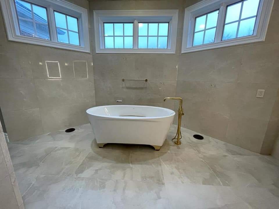 Bathroom with a white freestanding bathtub, gold faucet, and large gray tiles. Windows above.