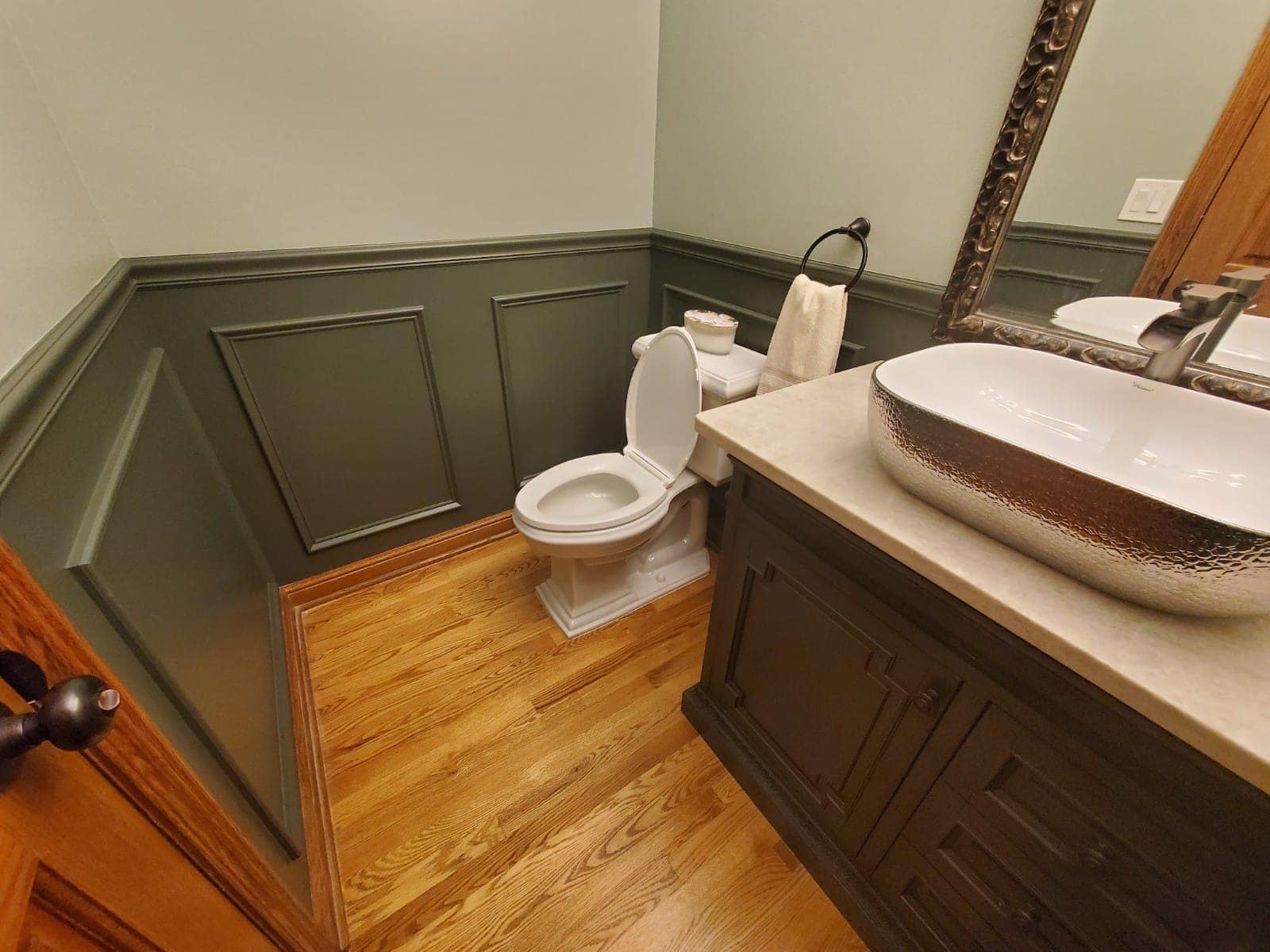 Small bathroom with green paneled walls, white toilet, wooden floor, and decorative sink.