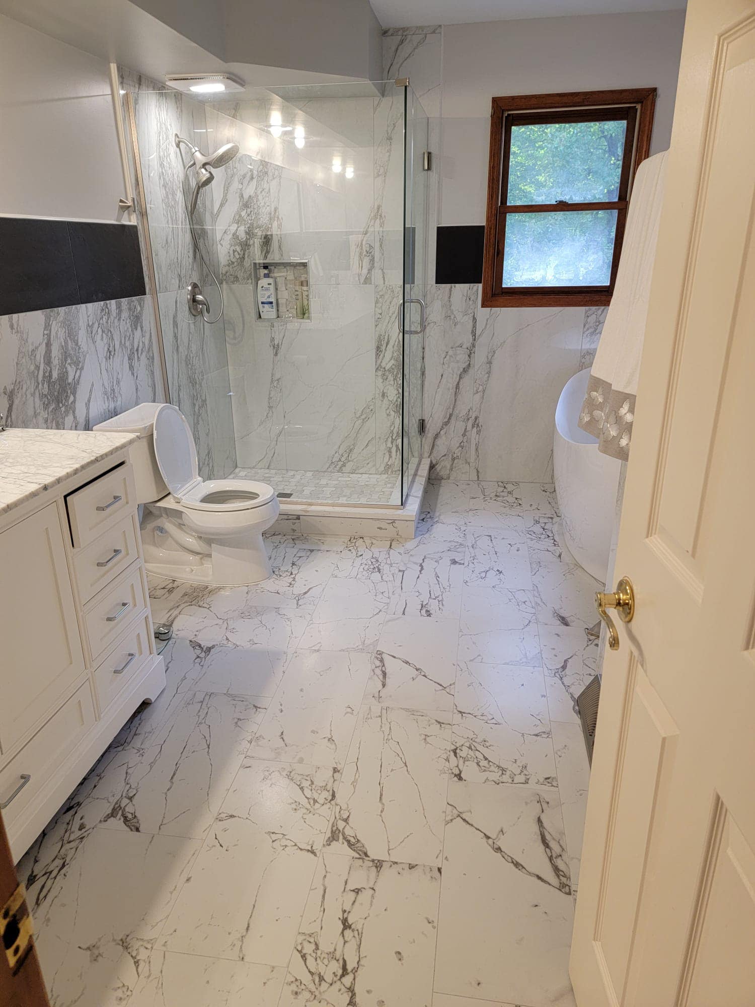 Bathroom with white marble tile, shower, toilet, vanity, and a window.