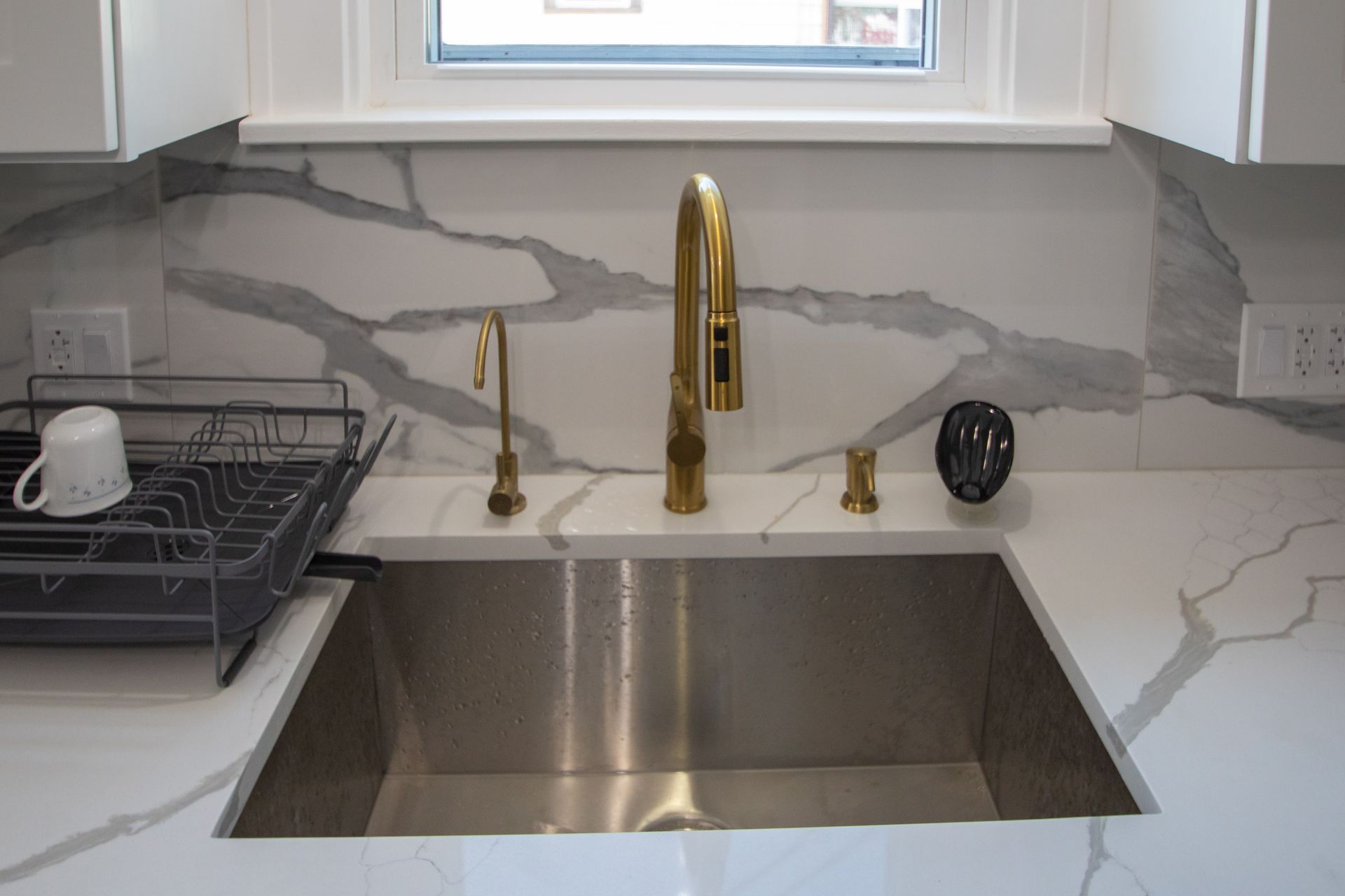 Stainless steel kitchen sink with gold faucet, set against marble backsplash. A dish rack sits on the left.
