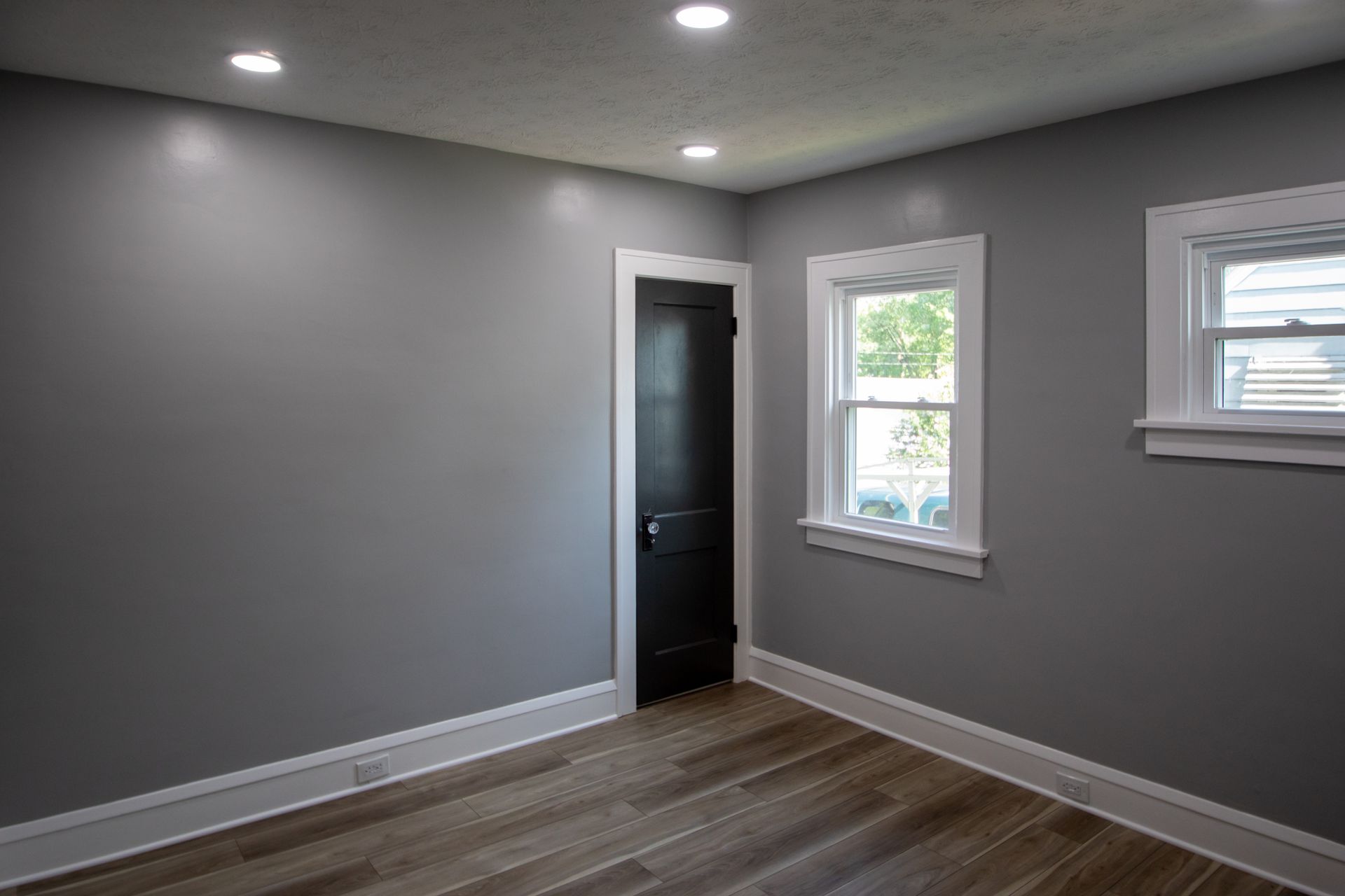 Empty room with gray walls, white trim, wood-look floor, black door, and two windows.