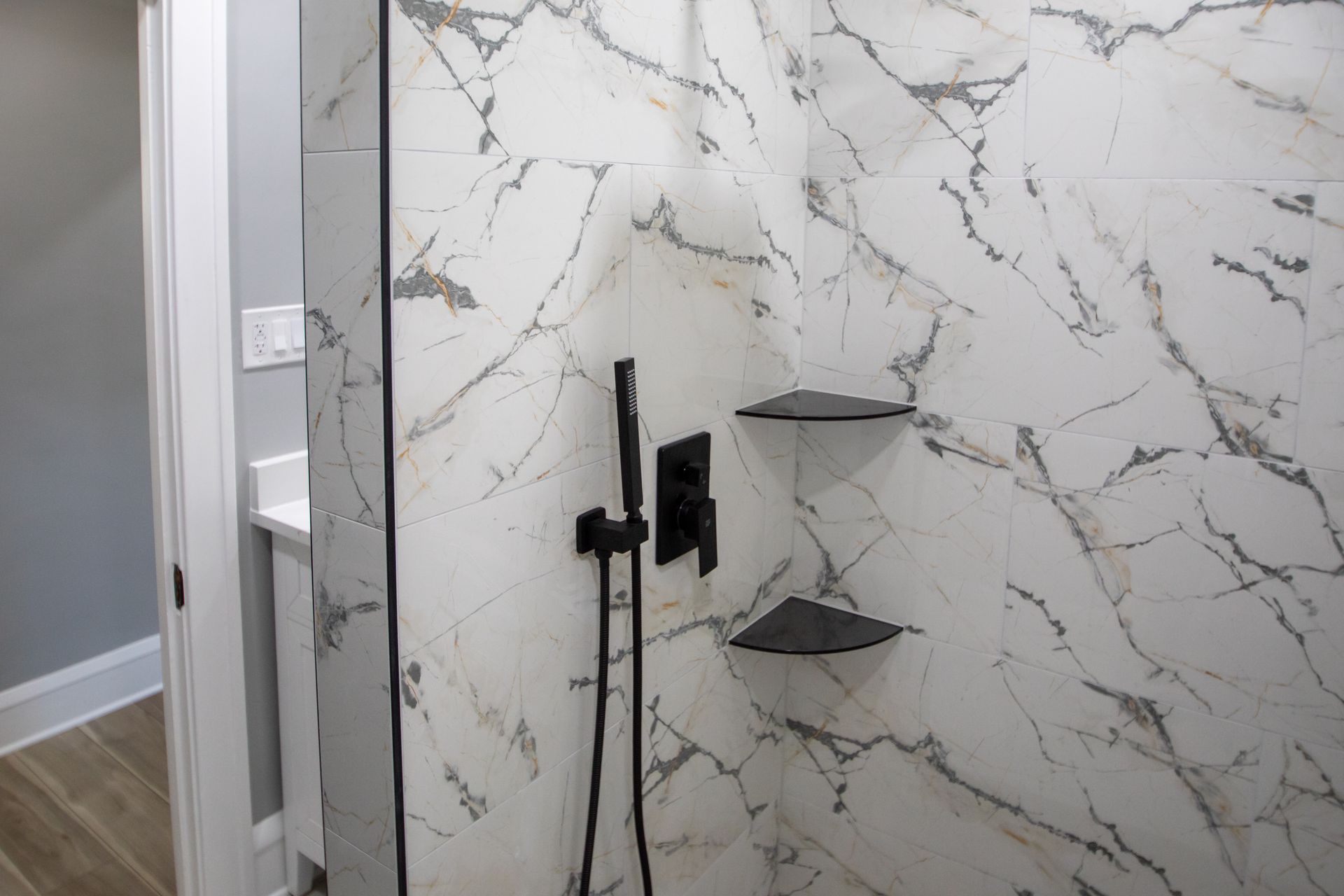Shower interior with white marble-patterned tile, black fixtures, and built-in black corner shelves.
