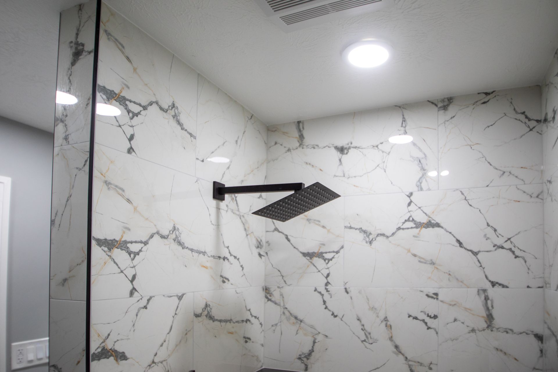 White marble tile shower with black shower head and overhead light.