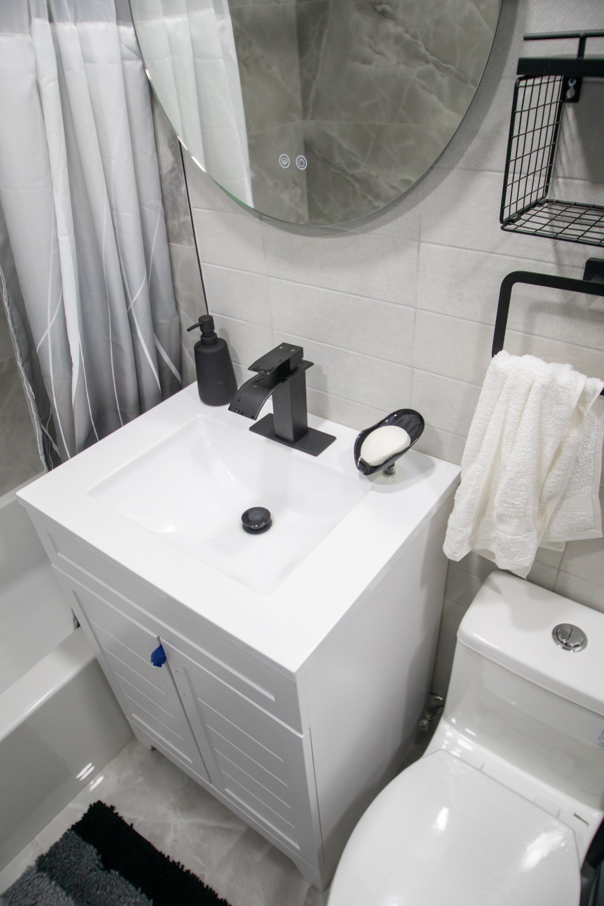 White bathroom vanity with black faucet, soap dish, and toilet. A round mirror hangs above.