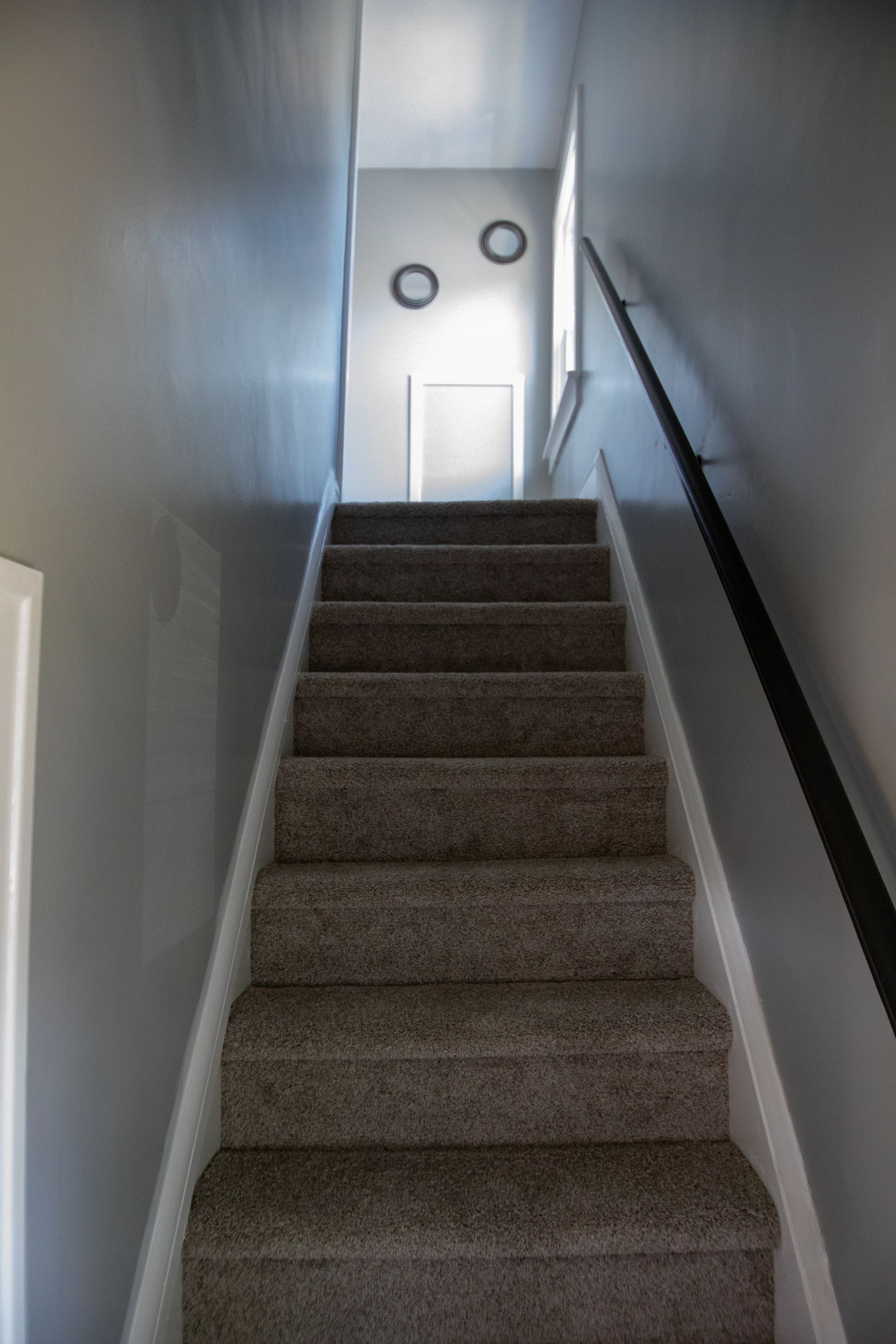 Staircase with gray carpet and walls, leading to a doorway, two round clocks, and a window.