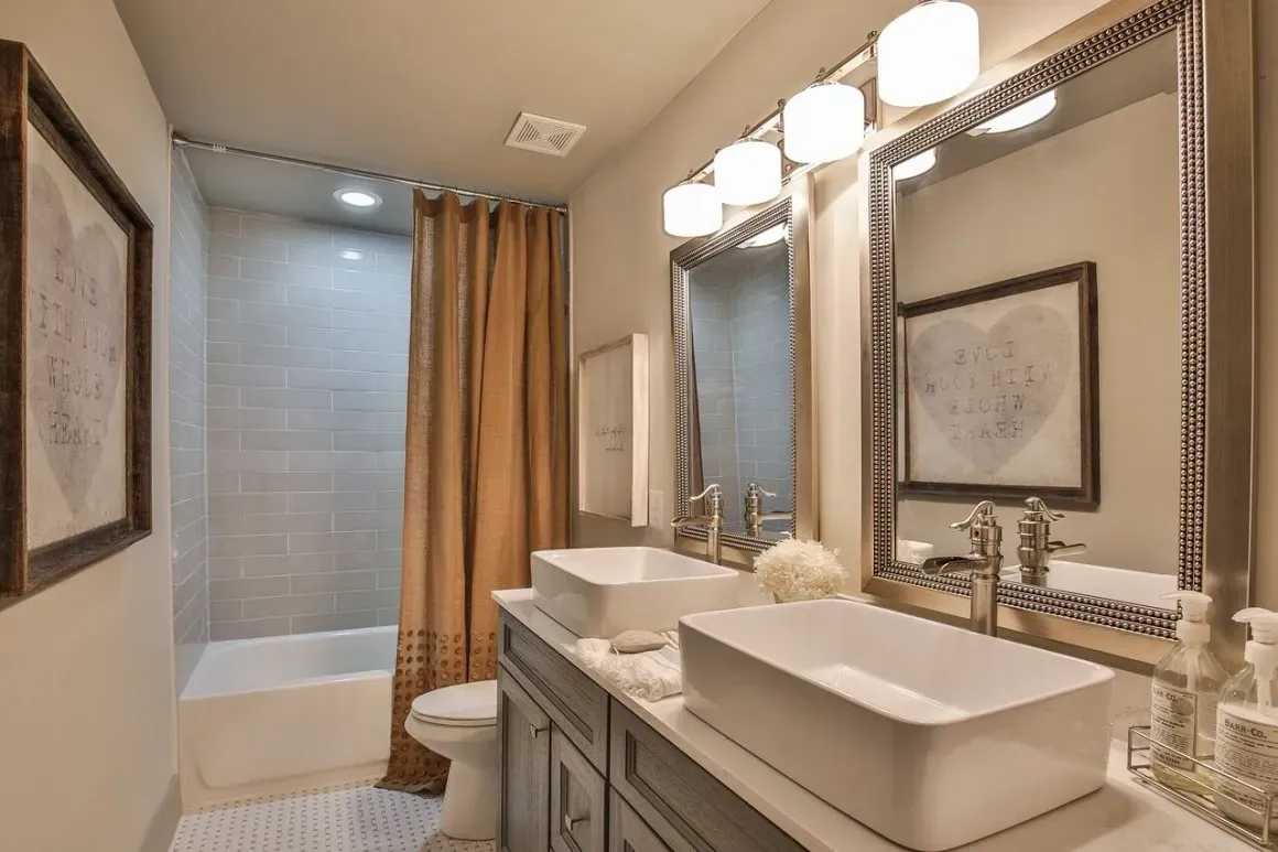 Bathroom with double sinks, a bathtub, and tan shower curtain. Gray cabinets and neutral walls.