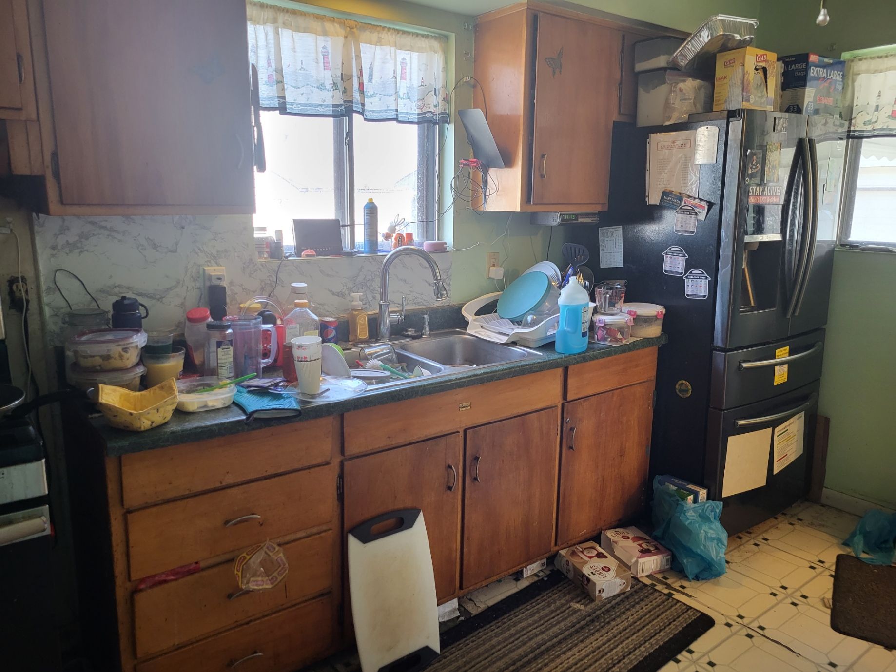 Messy kitchen with cluttered countertops and a full sink. Old wooden cabinets and a dark refrigerator.