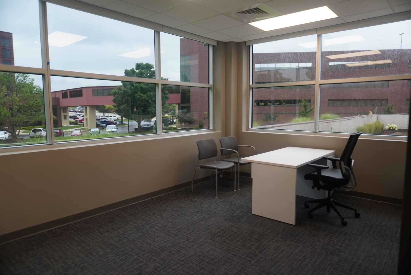 An empty office with large windows overlooking an exterior parking lot. Two chairs and a desk sit inside.