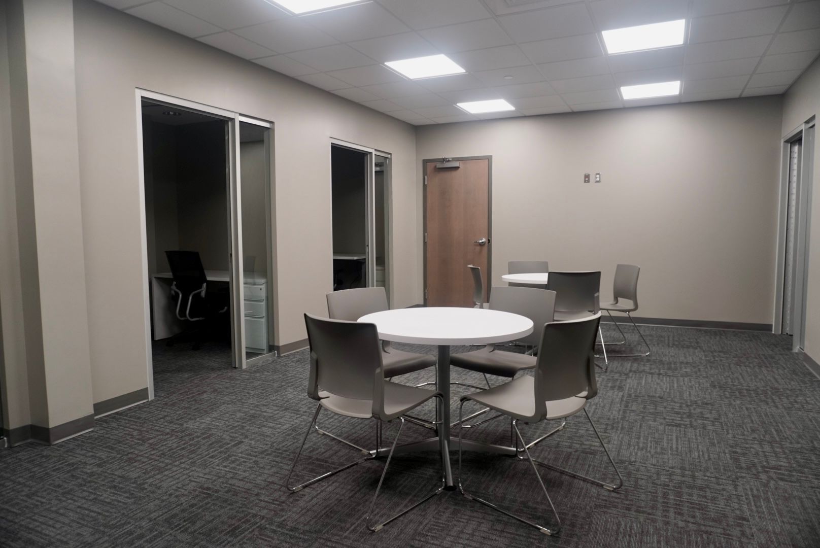 Room with a round table, chairs, office cubicle visible, and a closed door. Carpet flooring and neutral walls.