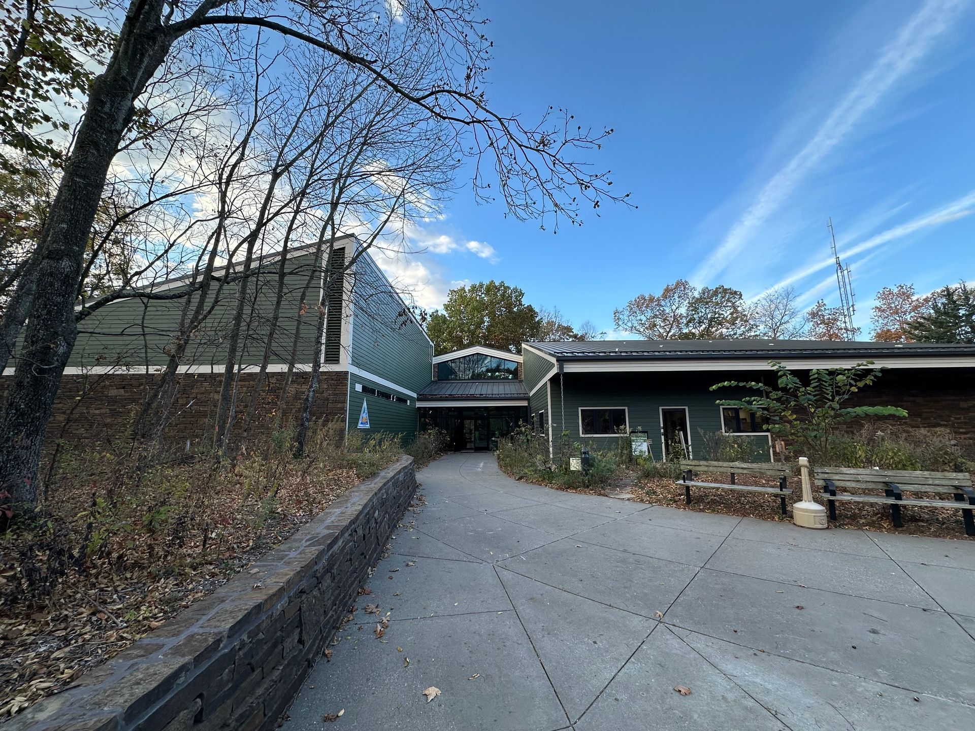 Building with angled roof, green siding, brick base, paved path, trees. Blue sky with cloud streaks.