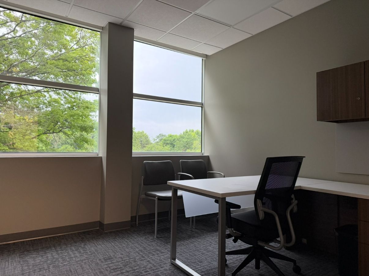 Empty office with desk, chair, two chairs, and window with a view of trees.