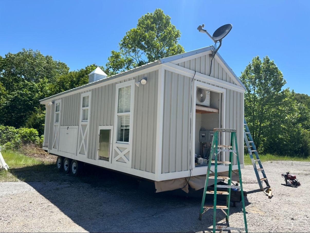 Light gray tiny house on a trailer with open door, ladder, satellite dish, and surrounding trees.