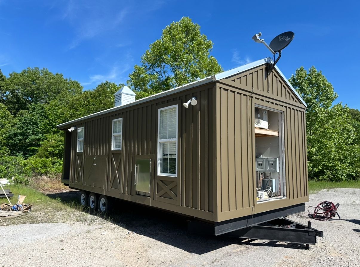 A mobile, olive-green tiny home on a trailer with a satellite dish, windows, and open doorway.