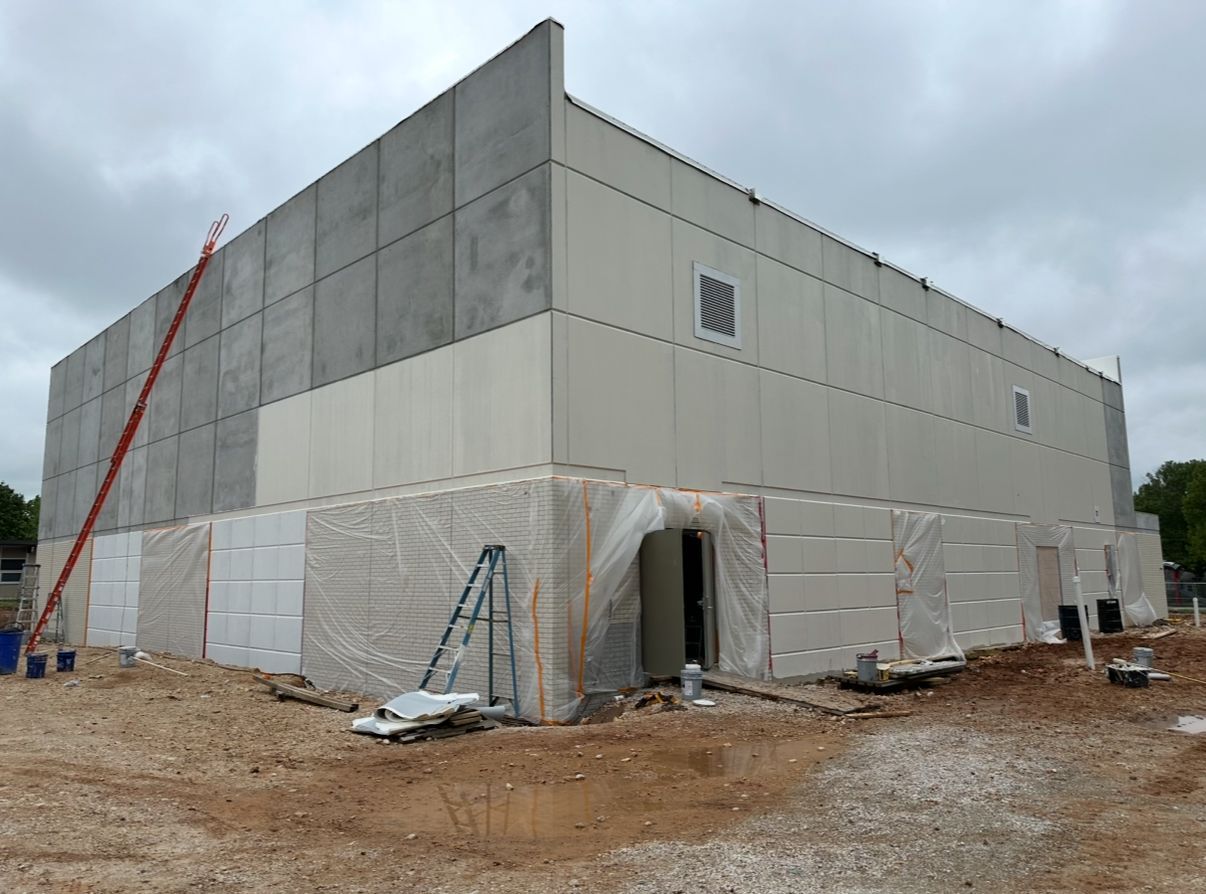 Construction site with a large building, exterior walls partly covered in gray and beige panels.