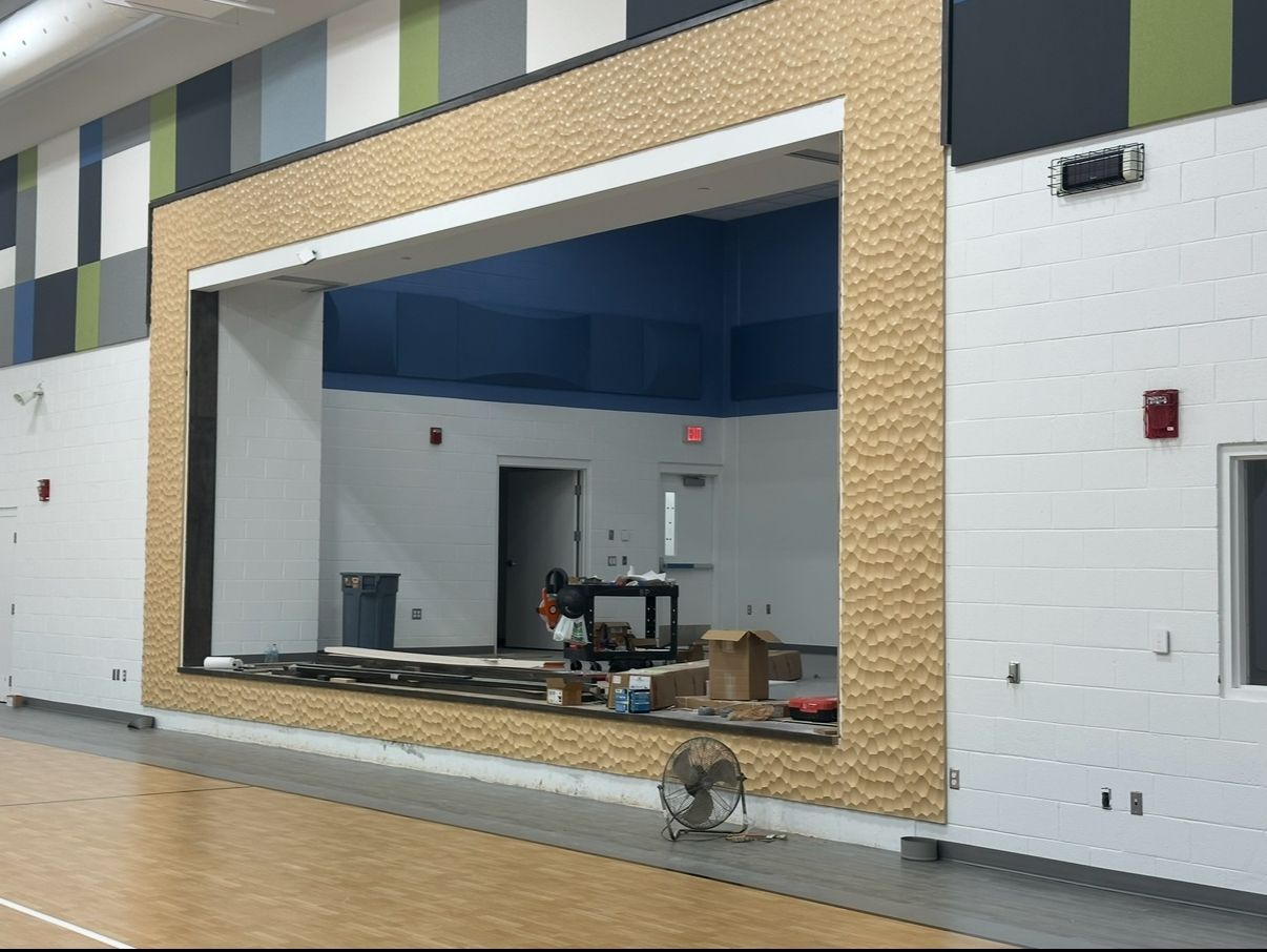 Gymnasium with stage under construction; light brown brick-like frame, blue ceiling, white walls, wooden floor.