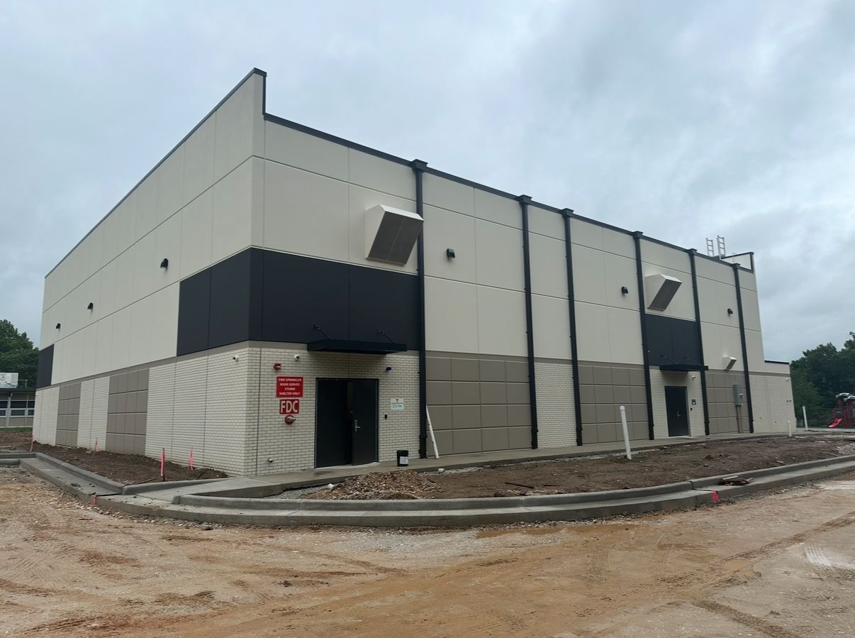 Exterior of a modern building under construction; tan and black facade; gray siding at the base; cloudy sky.