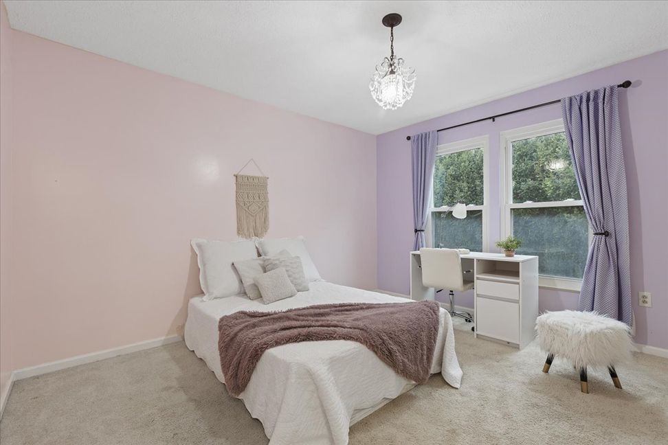 Bedroom with pink and purple walls, white desk, bed with purple blanket, and fluffy ottoman.