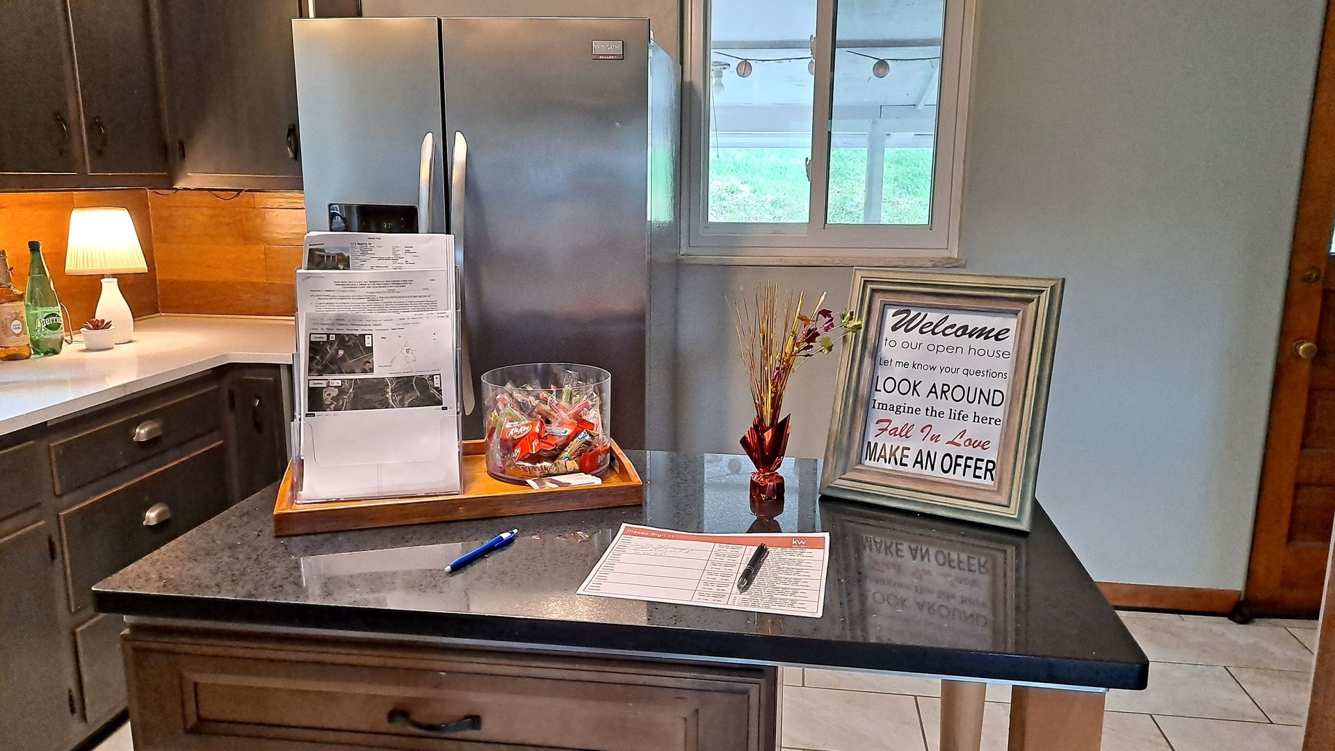 Kitchen island with stainless steel refrigerator, paperwork, and framed sign.