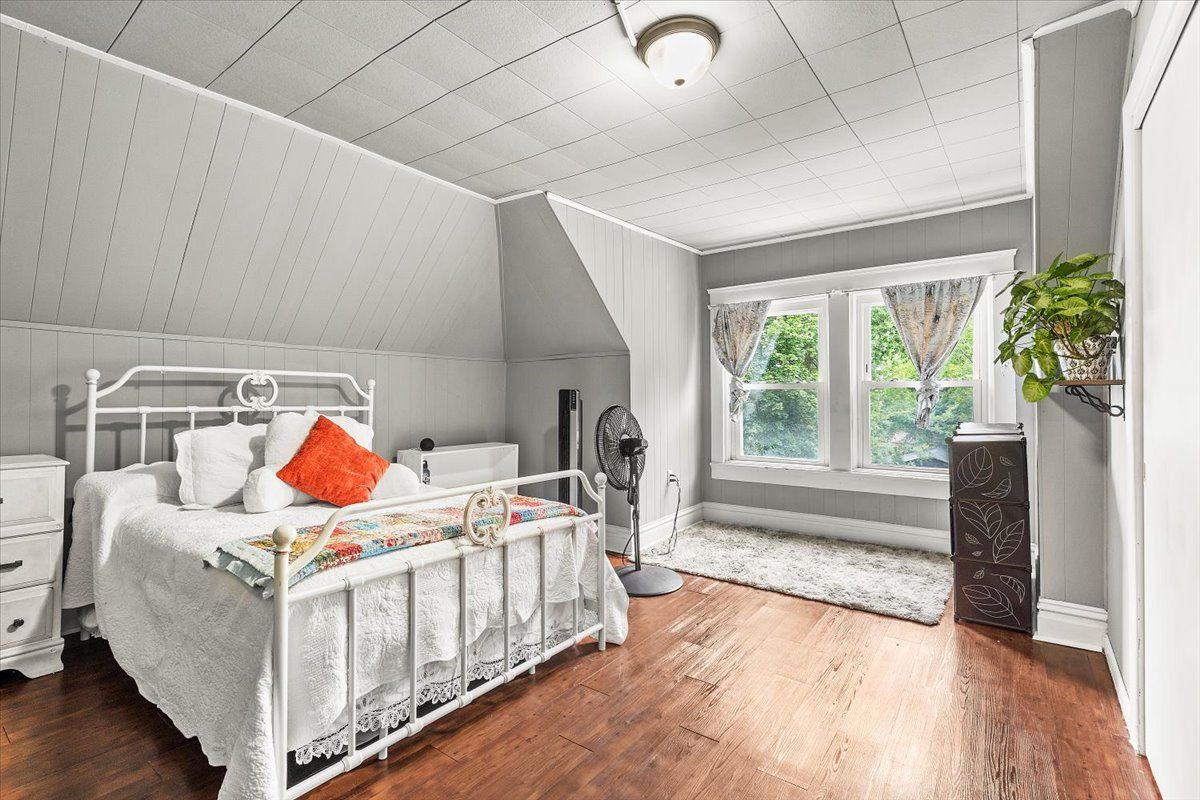 Bedroom with white bed, gray walls, and hardwood floor. Window with view, plant on the right.