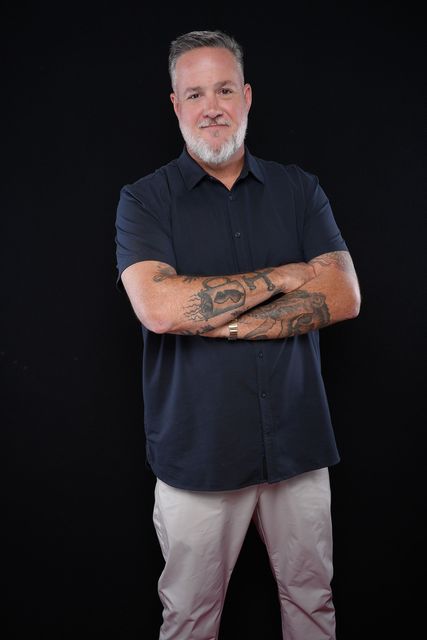 Man with shaved head, crossed arms, wearing a patterned button-down shirt and khakis, standing against a black background.