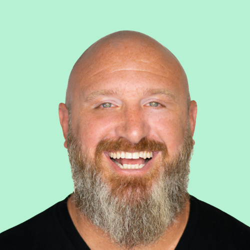 Bald man with a full beard smiles broadly, black shirt, light green background.